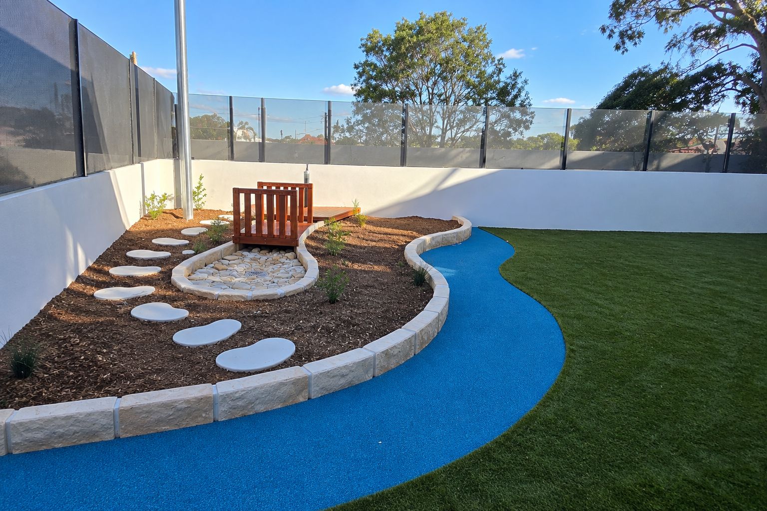 Outdoor playspace with artificial turf, a winding bright blue soft-fall path, a raised garden bed edged with stone blocks, white stepping stones and a small wooden bridge